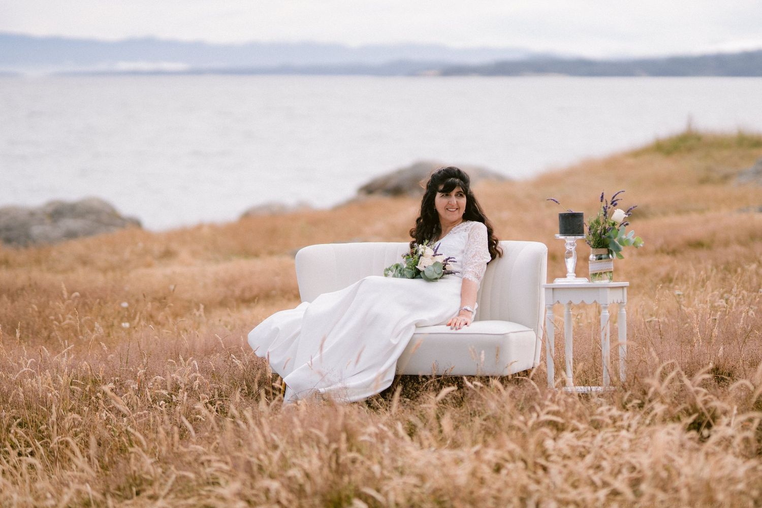 Bride in white gown sits on ivory sofa in golden field by the sea, holding bouquet.
