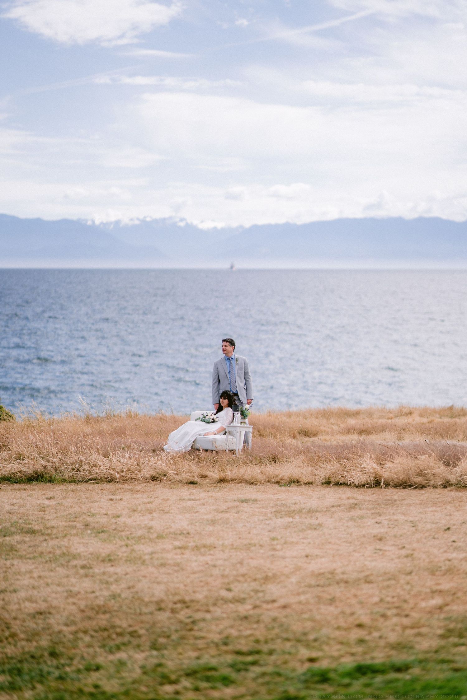 Bride and groom pose on a scenic coastal bluff with mountains and blue ocean waters in the background.
