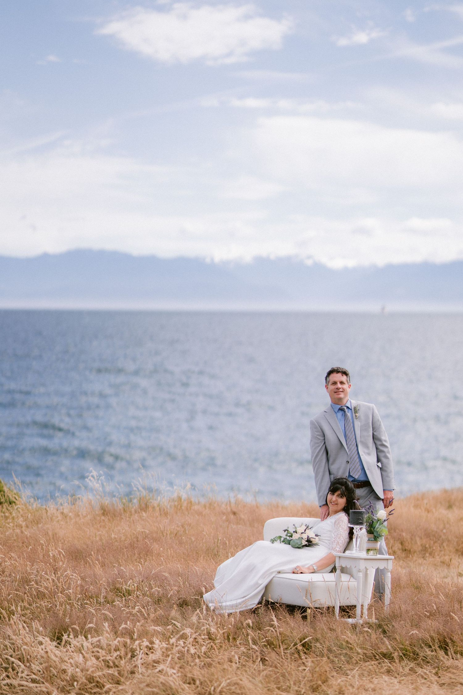 Bride and groom posing by the ocean in a field, woman seated on white sofa, man standing in gray suit.