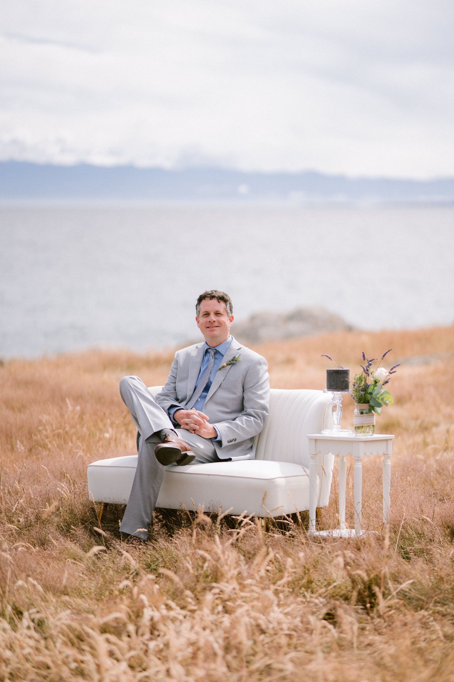 Groom in gray suit sitting on white sofa in golden field by the ocean.