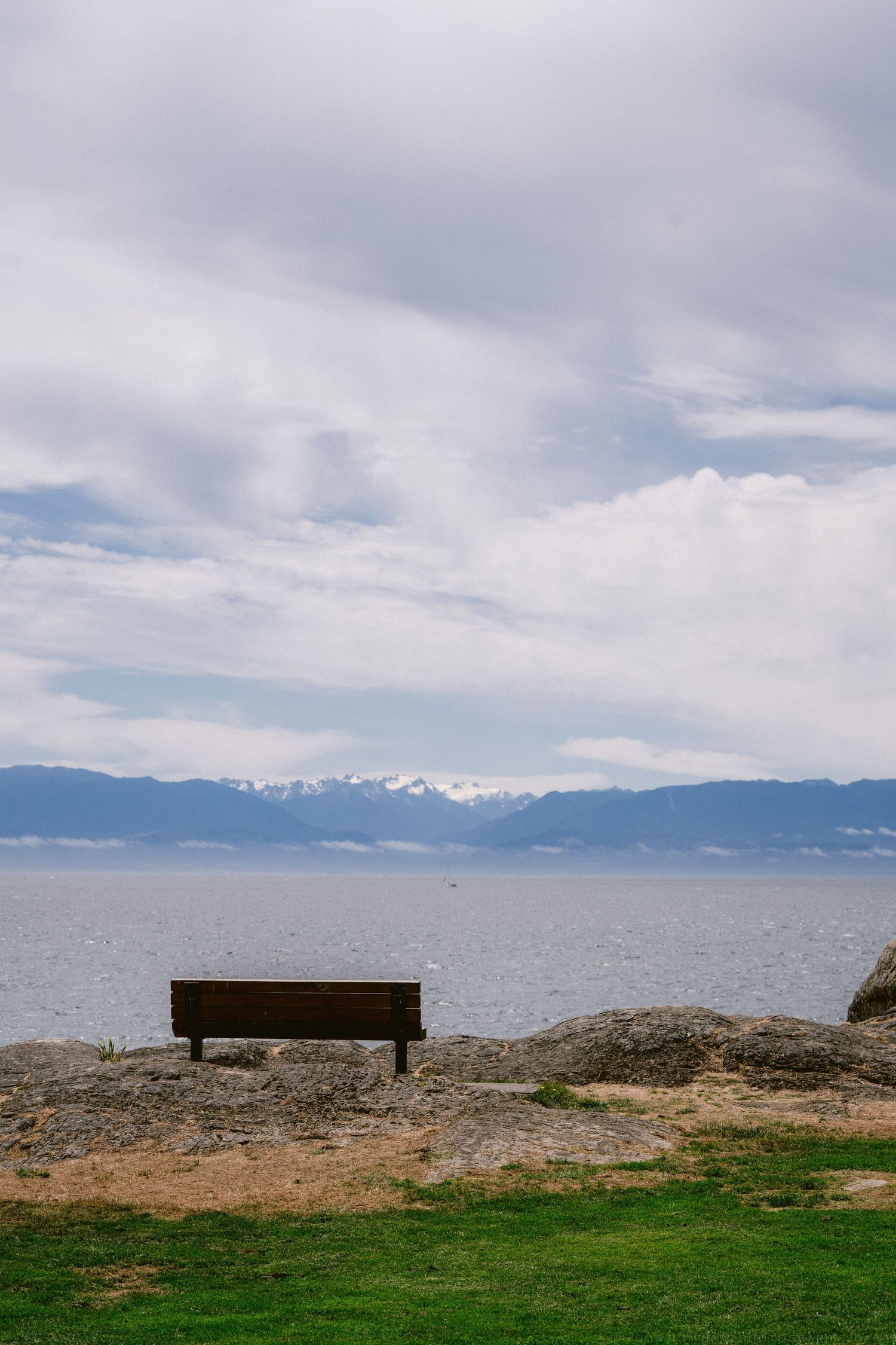 Lone wooden bench on rocky shore overlooking calm ocean with snow-capped mountains and cloudy sky.