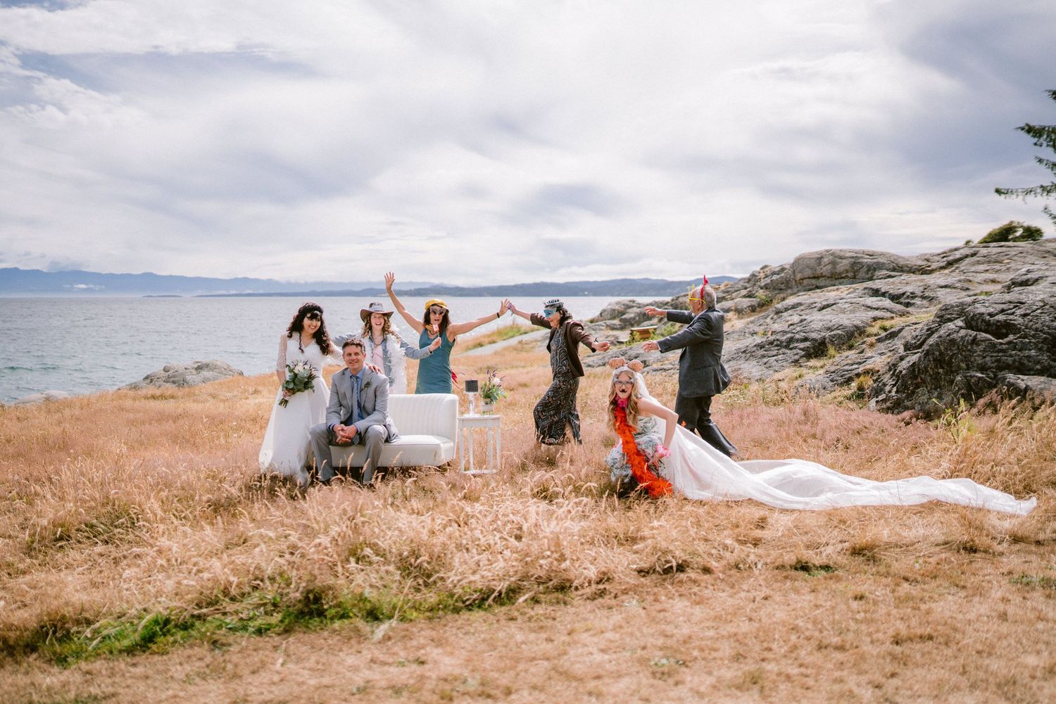 Wedding party celebrating outdoors near rocky coastline with bride's veil flowing in wind over golden grass.