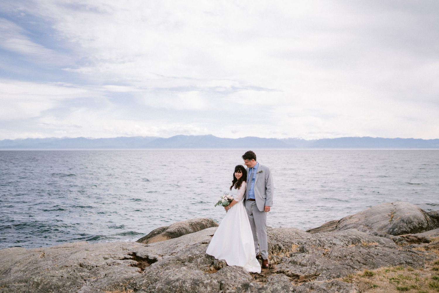 Bride and groom stand on rocky shoreline with mountain lake backdrop on their wedding day.