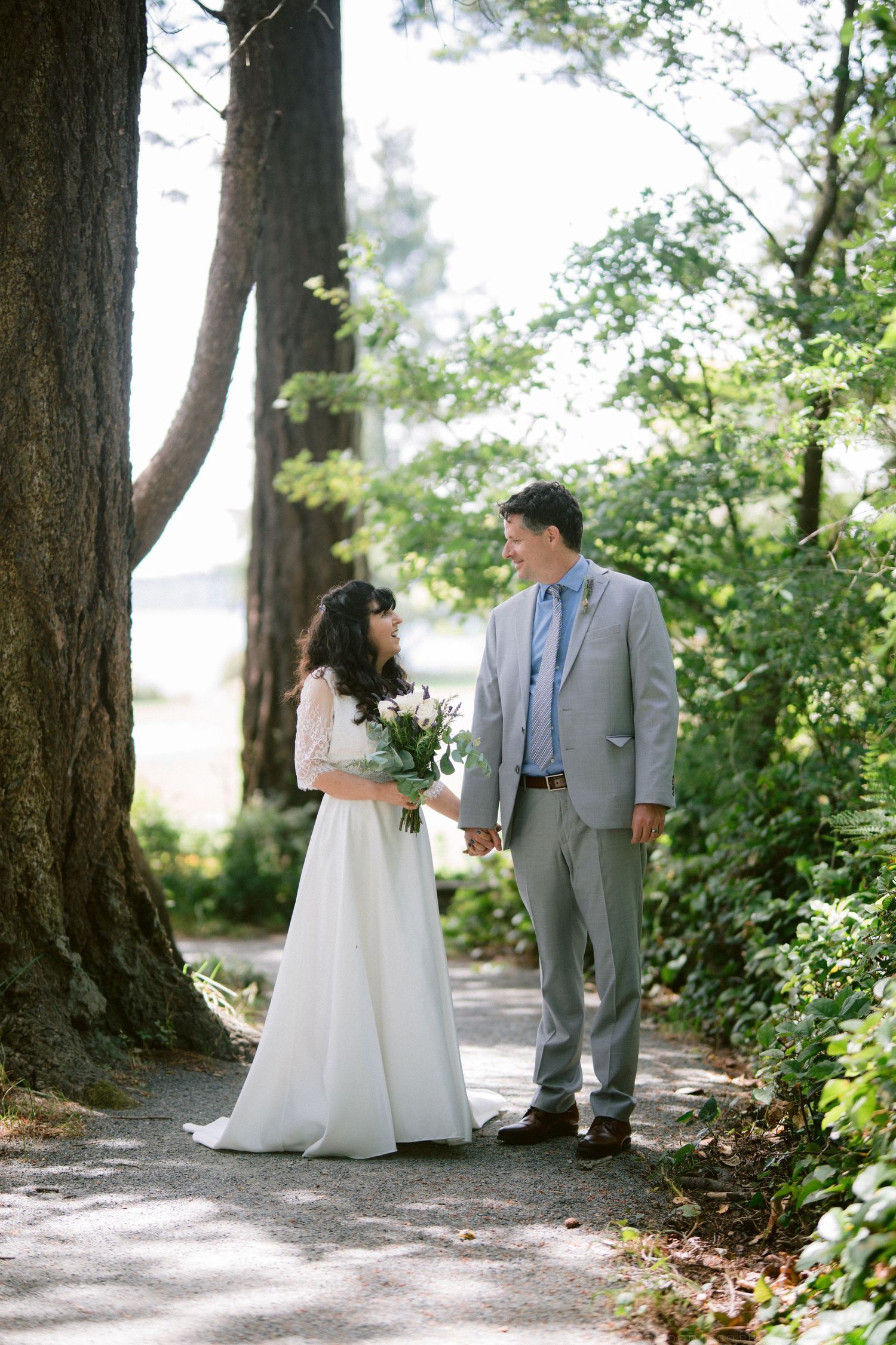 Bride and groom hold hands on tree-lined path near water on their wedding day.