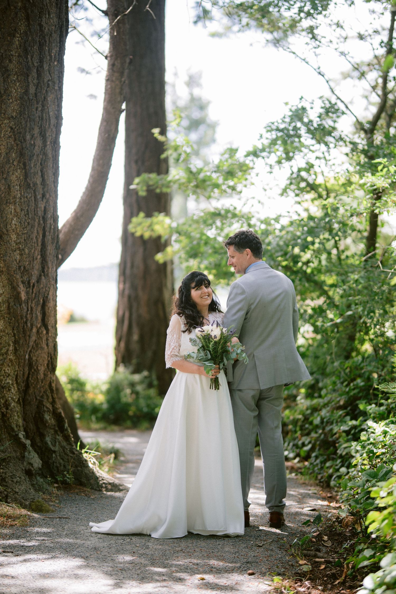 Bride and groom share a tender moment on a forest path surrounded by tall trees near water.