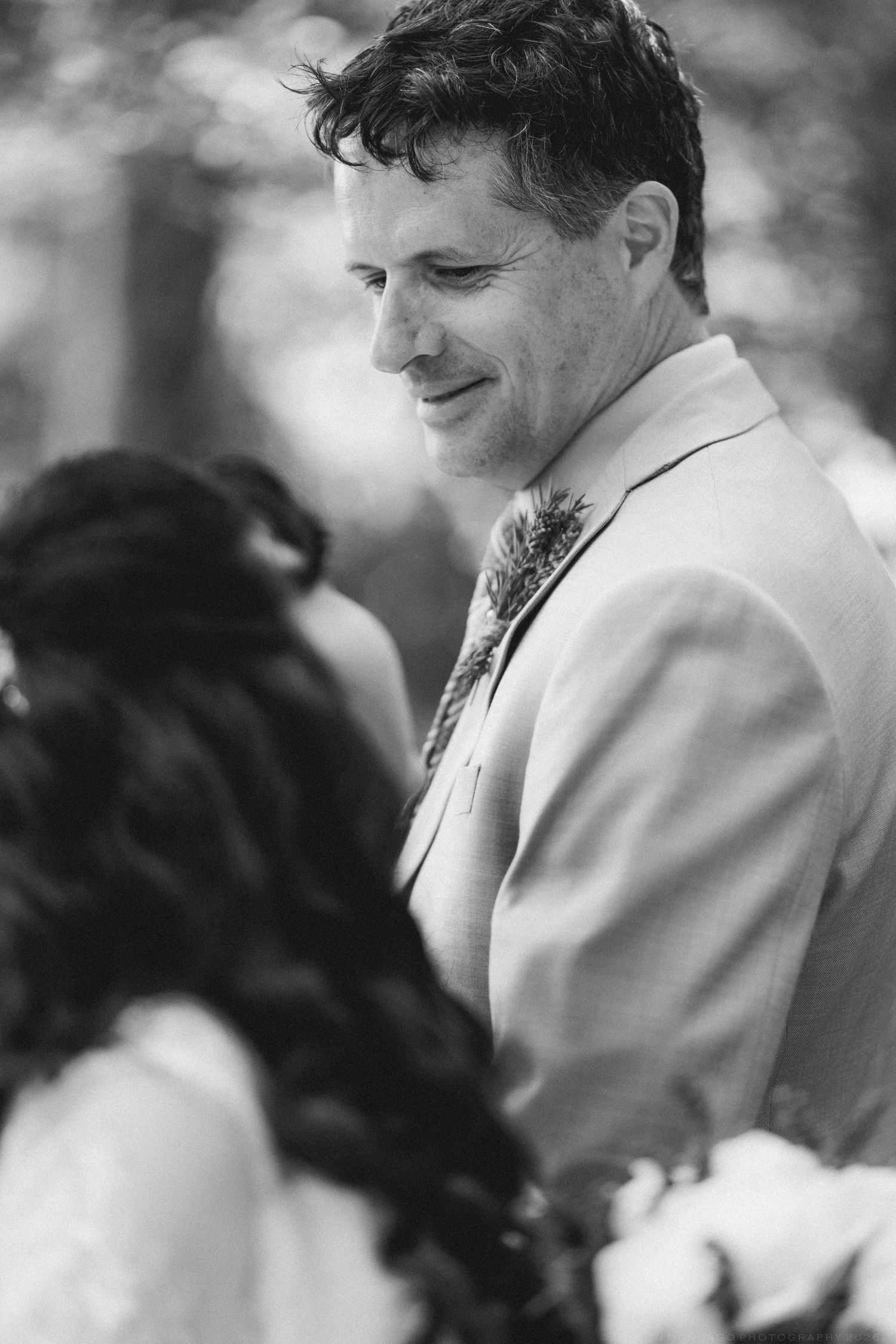 Black and white portrait of smiling groom in suit looking down at bride during wedding ceremony.