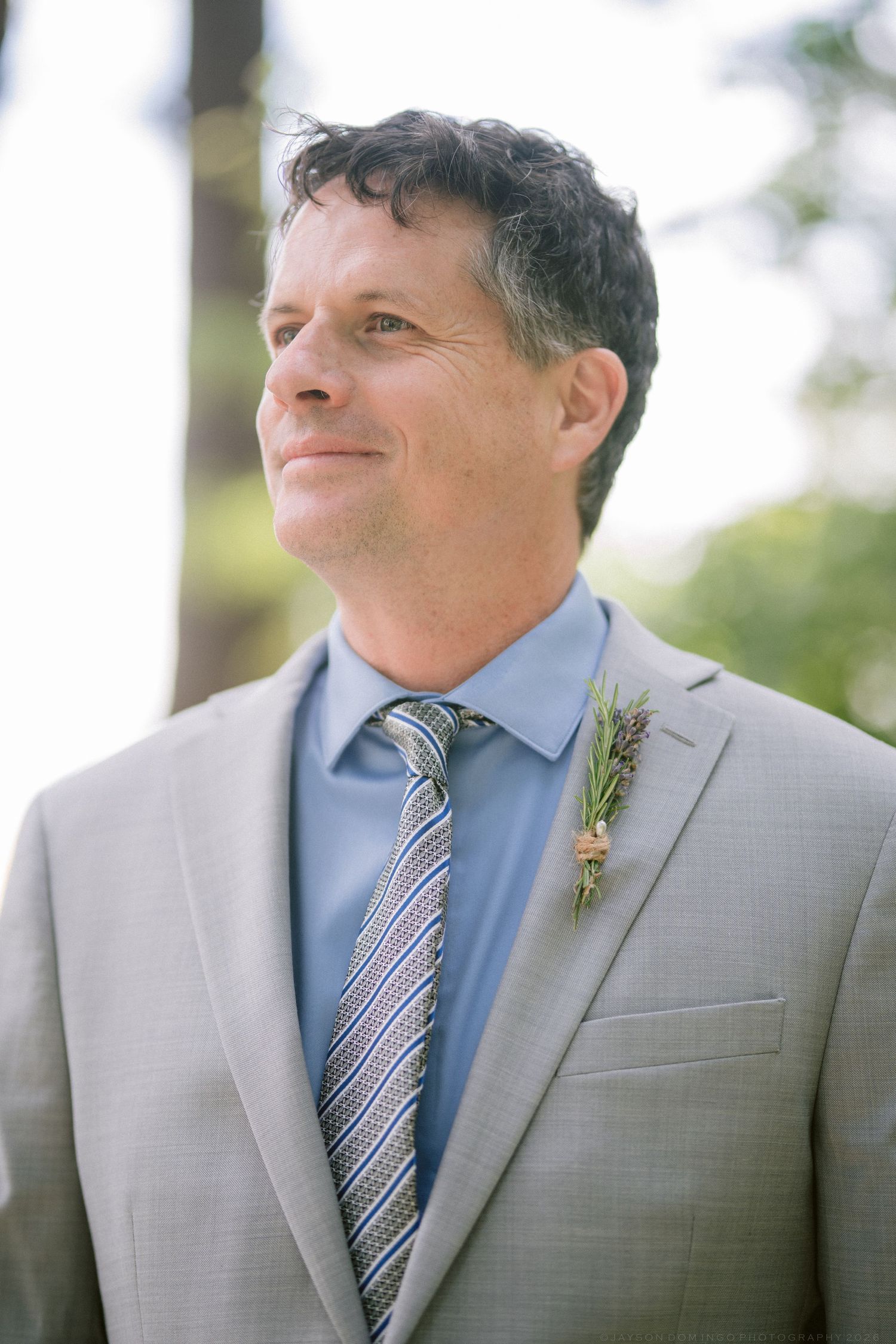 Groom in grey suit and striped tie with boutonniere smiling outdoors at a wedding.