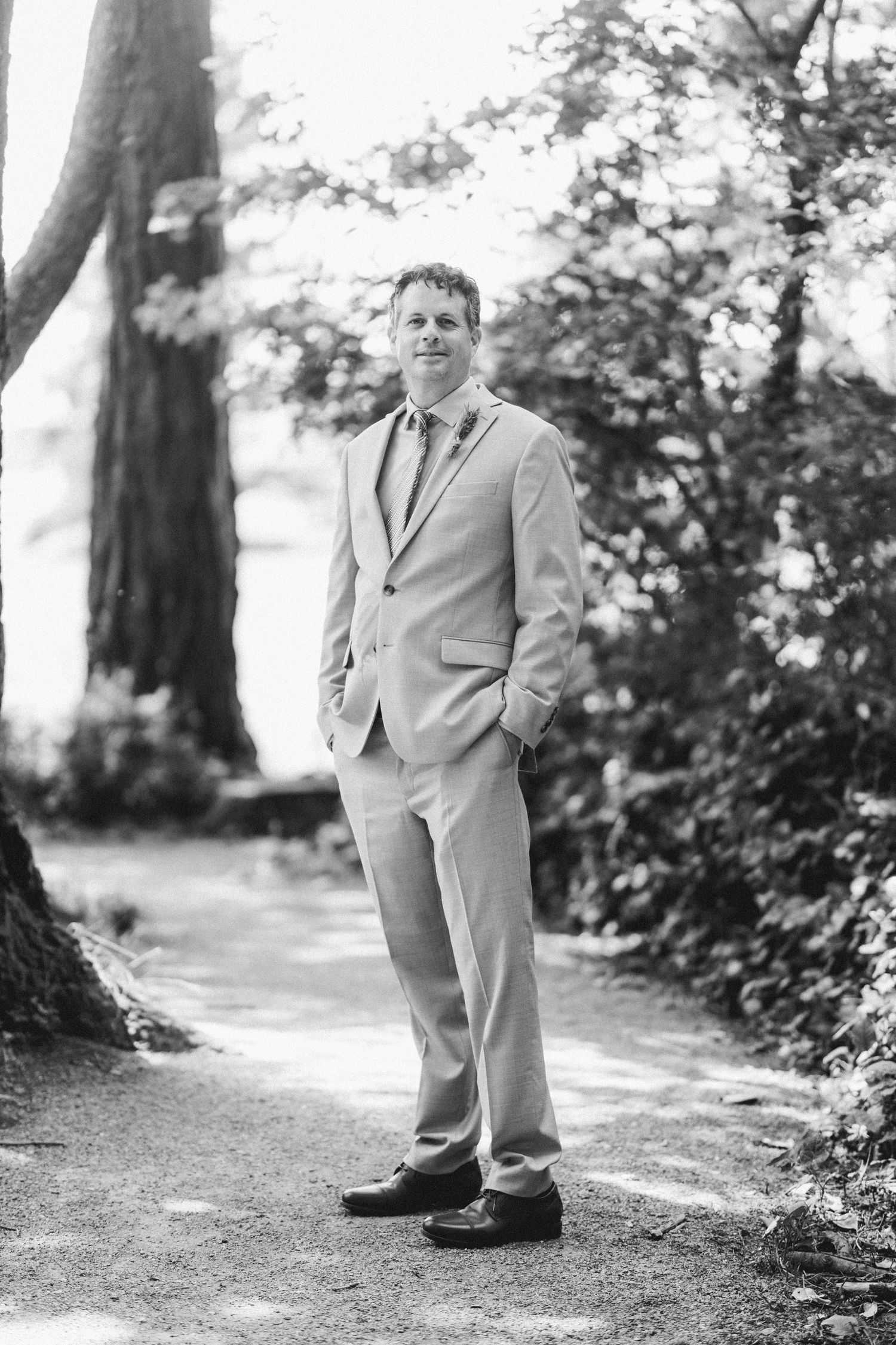 Black and white portrait of groom in light gray suit standing on a tree-lined garden path.