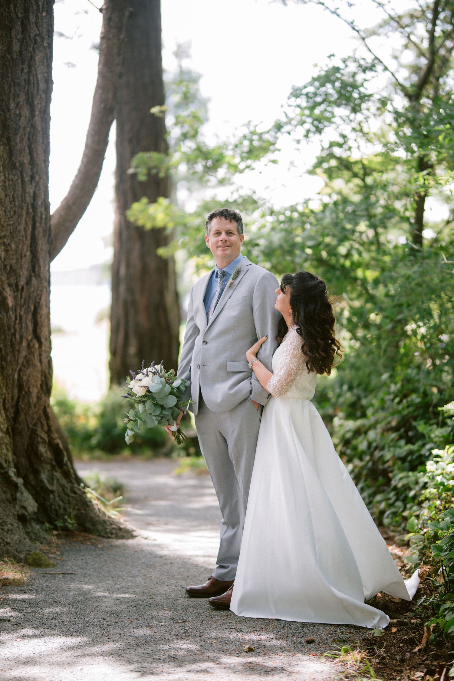 A groom in a gray suit holds a bouquet as his bride in a white gown embraces him on a tree-lined path.