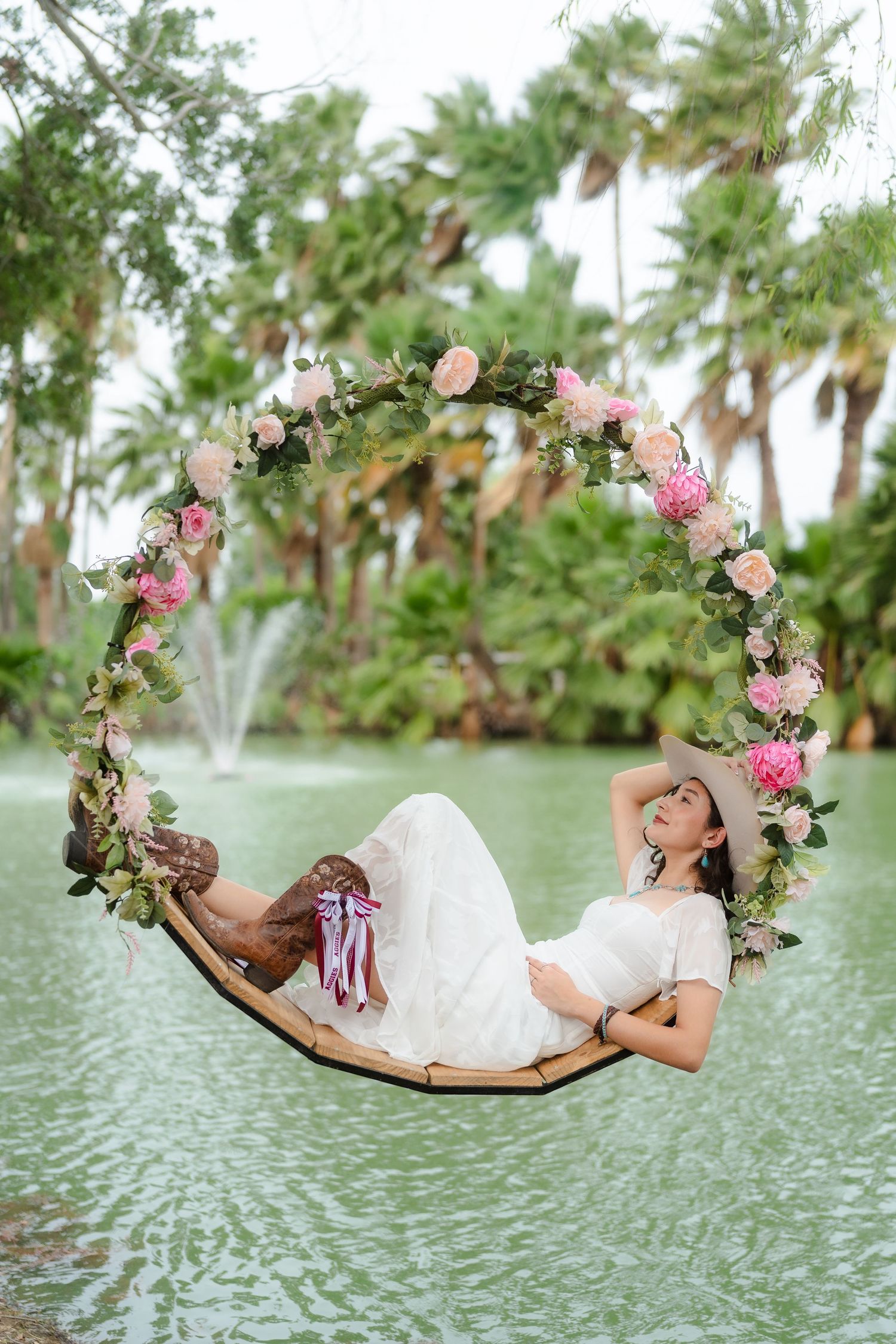Bride in white dress and hat reclines on a floral hoop swing over water, surrounded by tropical palms and a fountain.