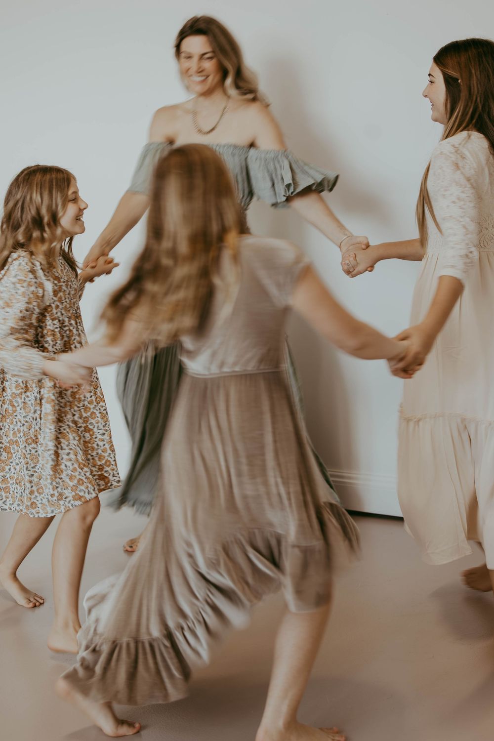 Four women in flowing dresses joyfully dance in a circle together in a bright, airy studio space.