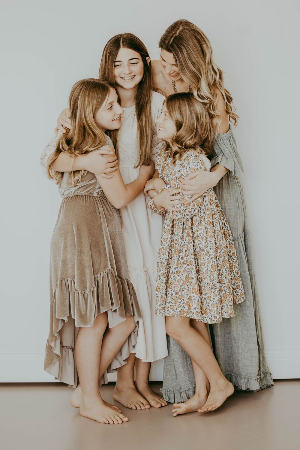 A mother and three daughters in flowing dresses share a warm group hug against a light background.