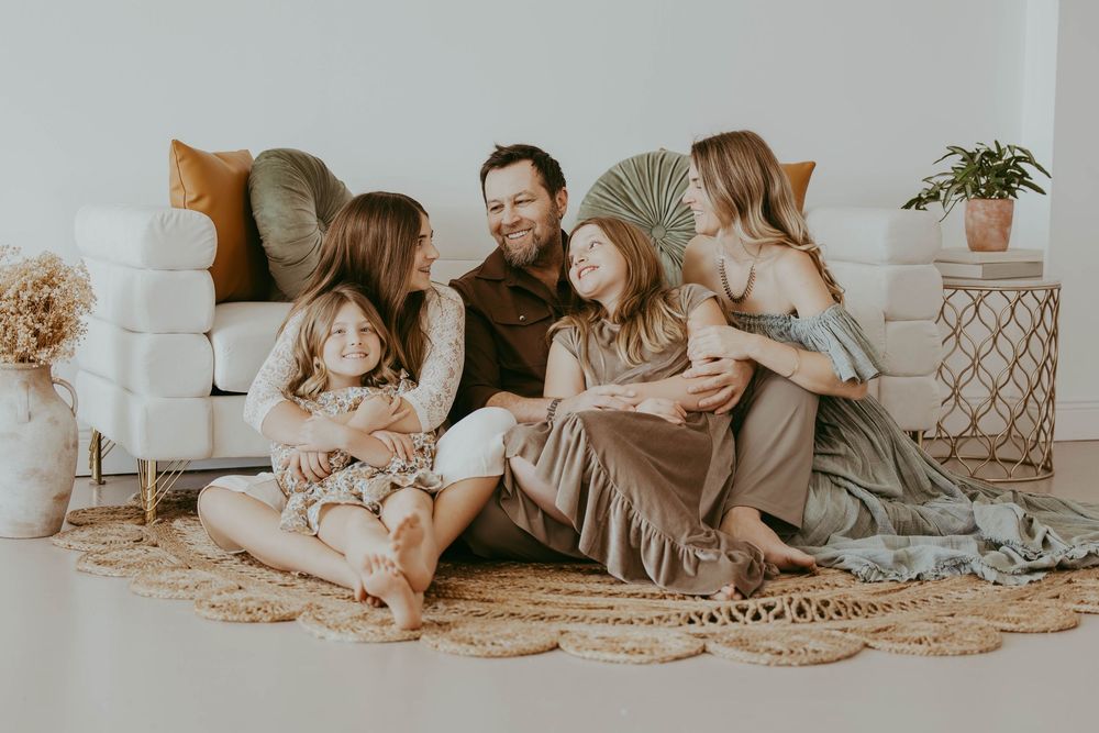 Happy family of five sitting together on a boho rug, smiling and laughing in a bright, cozy living room.