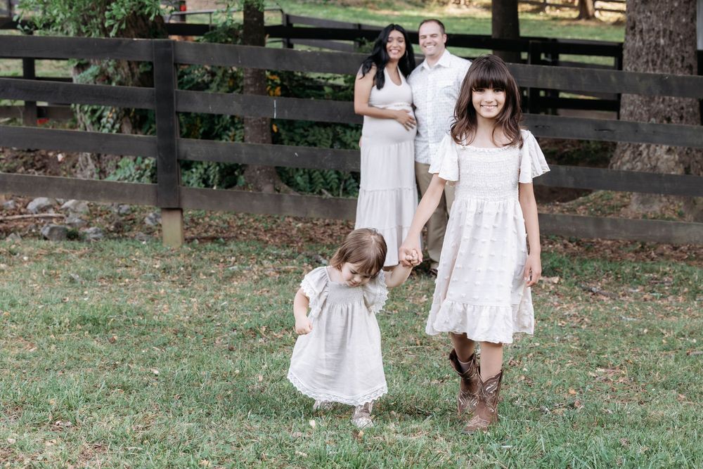 Family of four in white dresses poses outdoors near wooden fence on green grass.