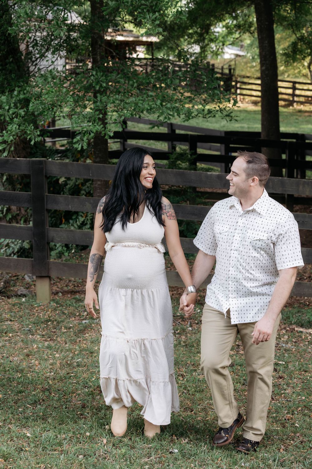 Pregnant woman in white dress holds hands with man in white shirt, walking and smiling outdoors near wooden fence.