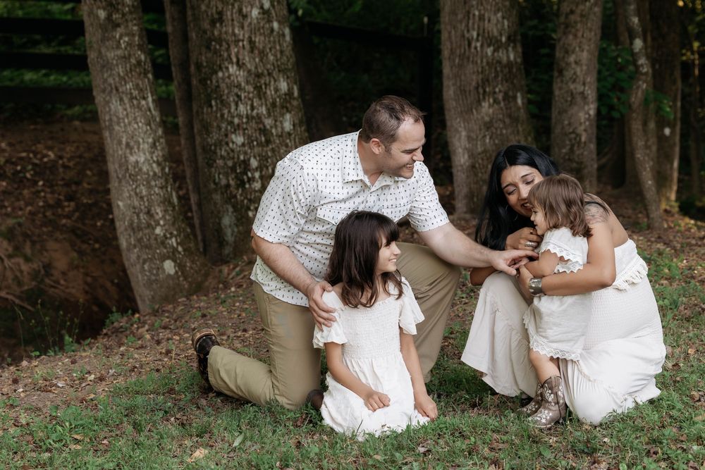 Family of four sharing a tender moment outdoors near trees, parents engaging with two young girls in white dresses.