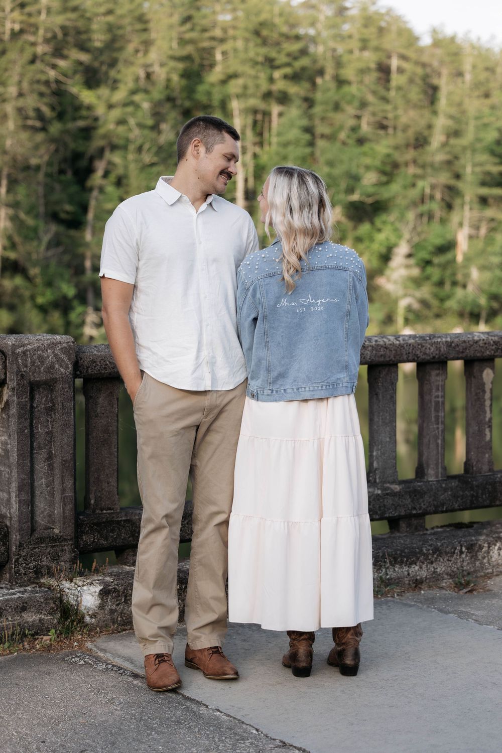 Couple standing on a bridge smiling at each other, woman wearing denim jacket and white maxi skirt, man in white polo and khakis.