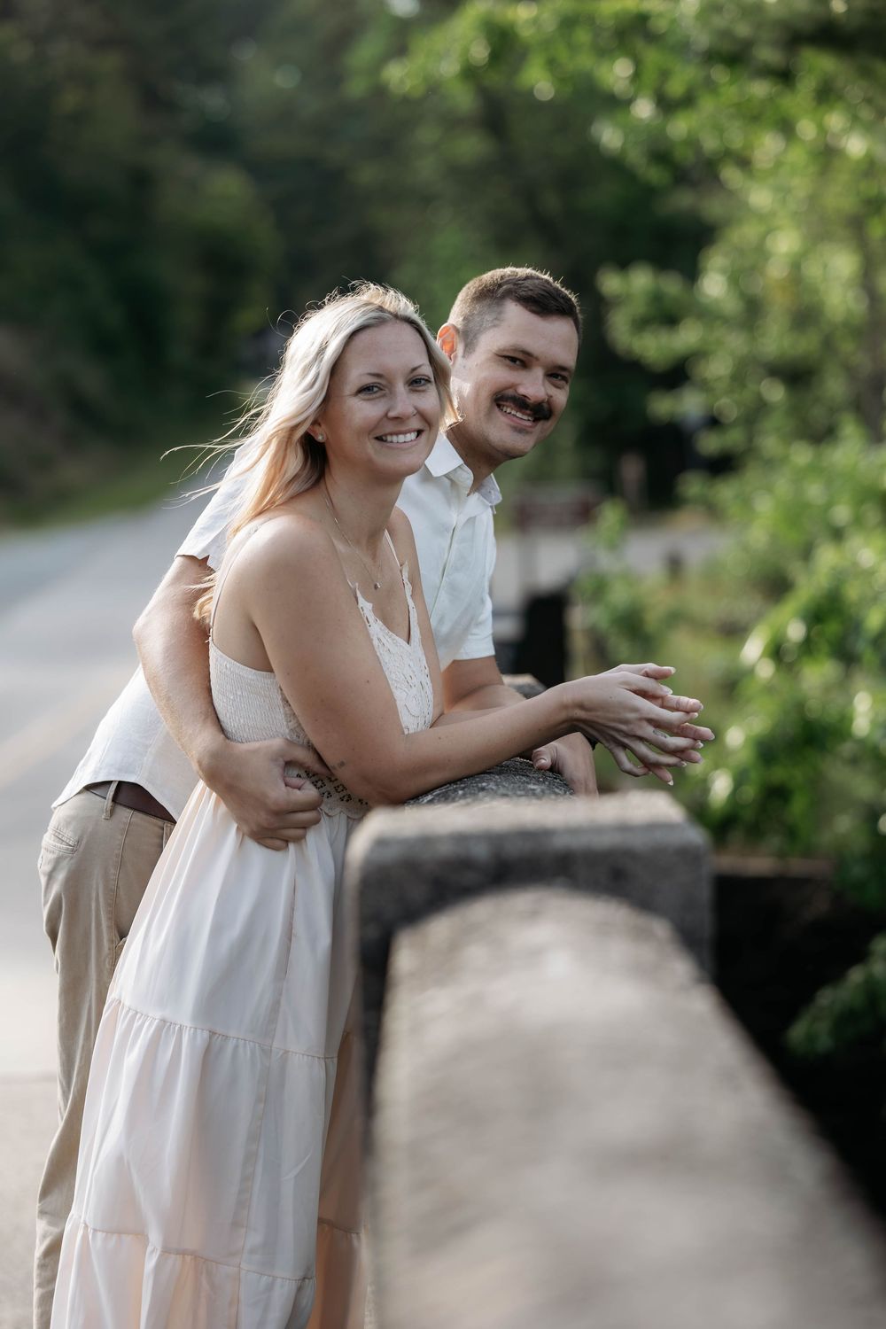 Happy couple embracing on a stone bridge surrounded by lush green trees, woman in white dress smiling outdoors.