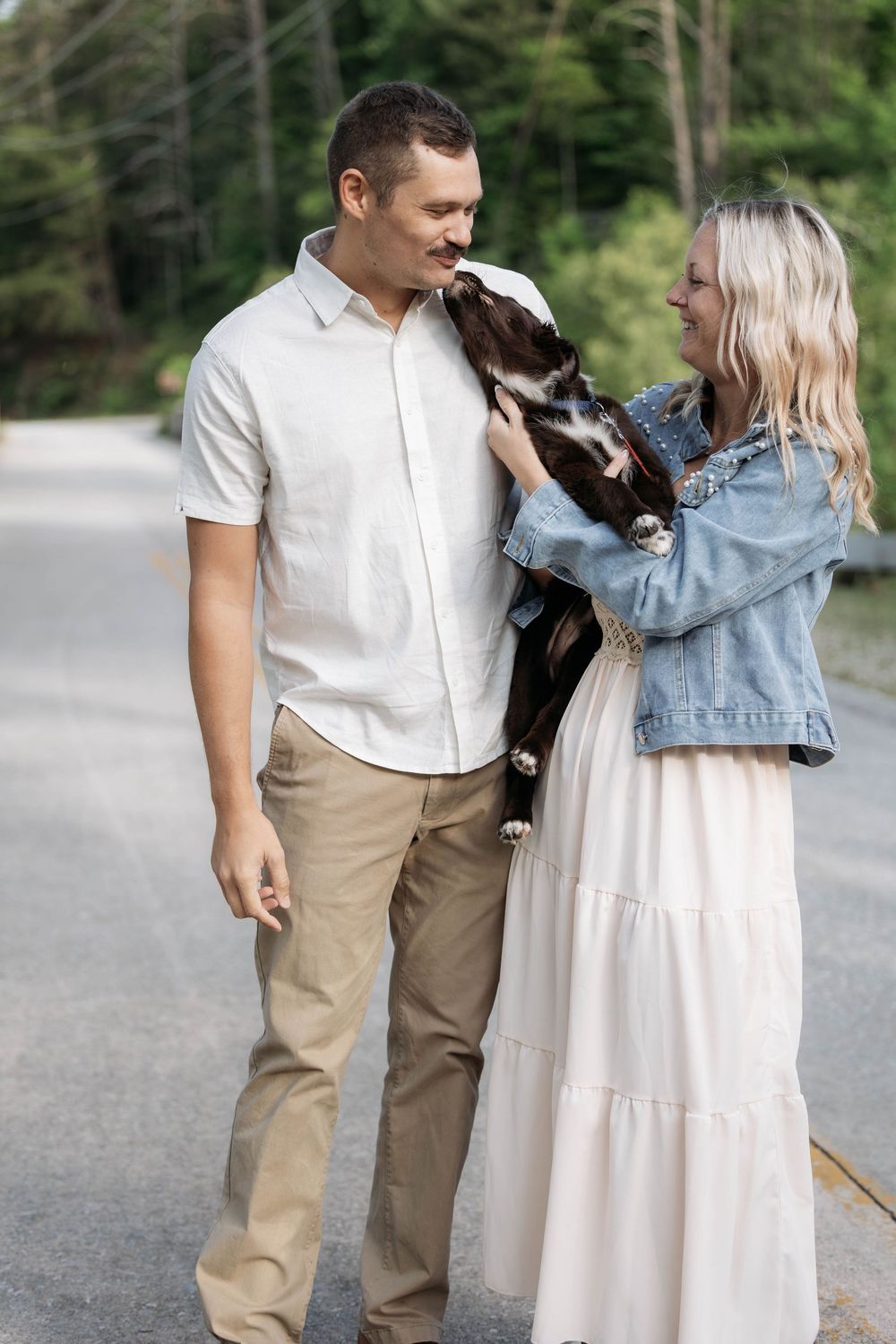 A couple smiles while holding a dog outdoors on a road surrounded by lush green trees.
