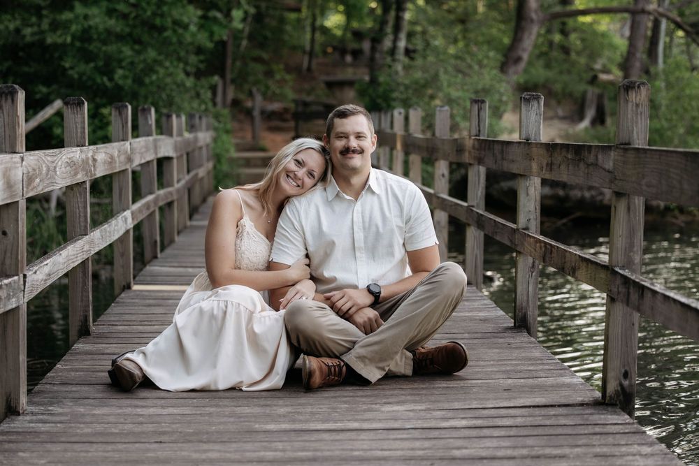Couple sitting on wooden bridge by water, woman in white dress leaning on man in white shirt, surrounded by trees.