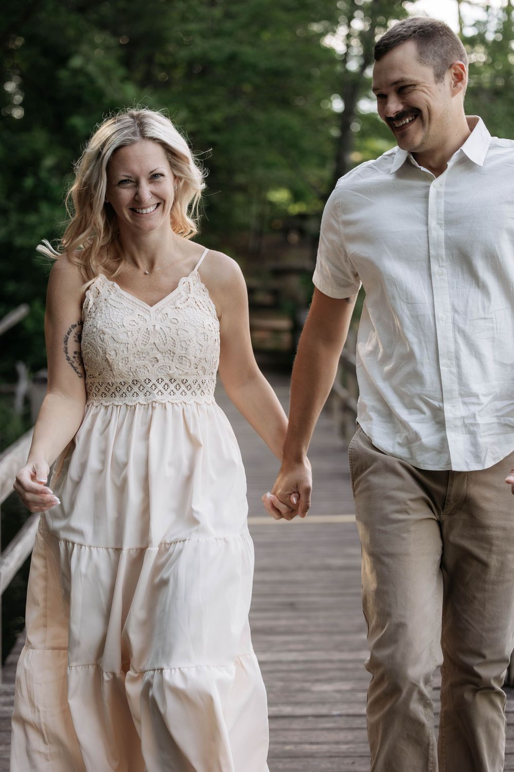 A smiling couple holds hands while walking on a wooden bridge surrounded by lush green trees.