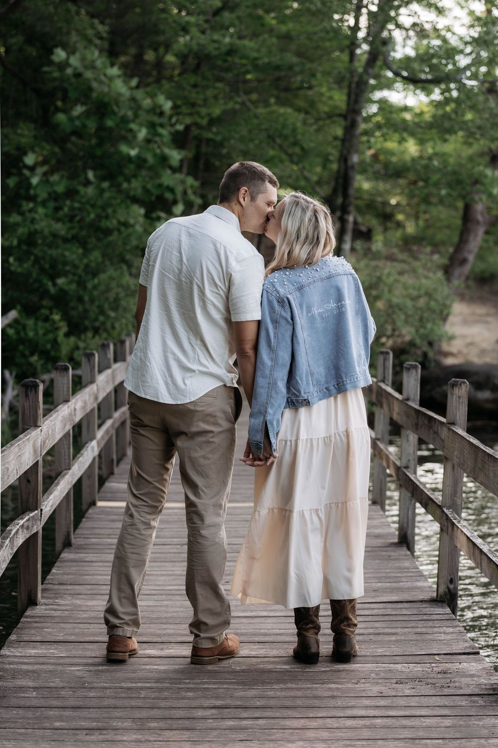 Couple kissing on a wooden bridge surrounded by lush green trees, woman in white dress and denim jacket.