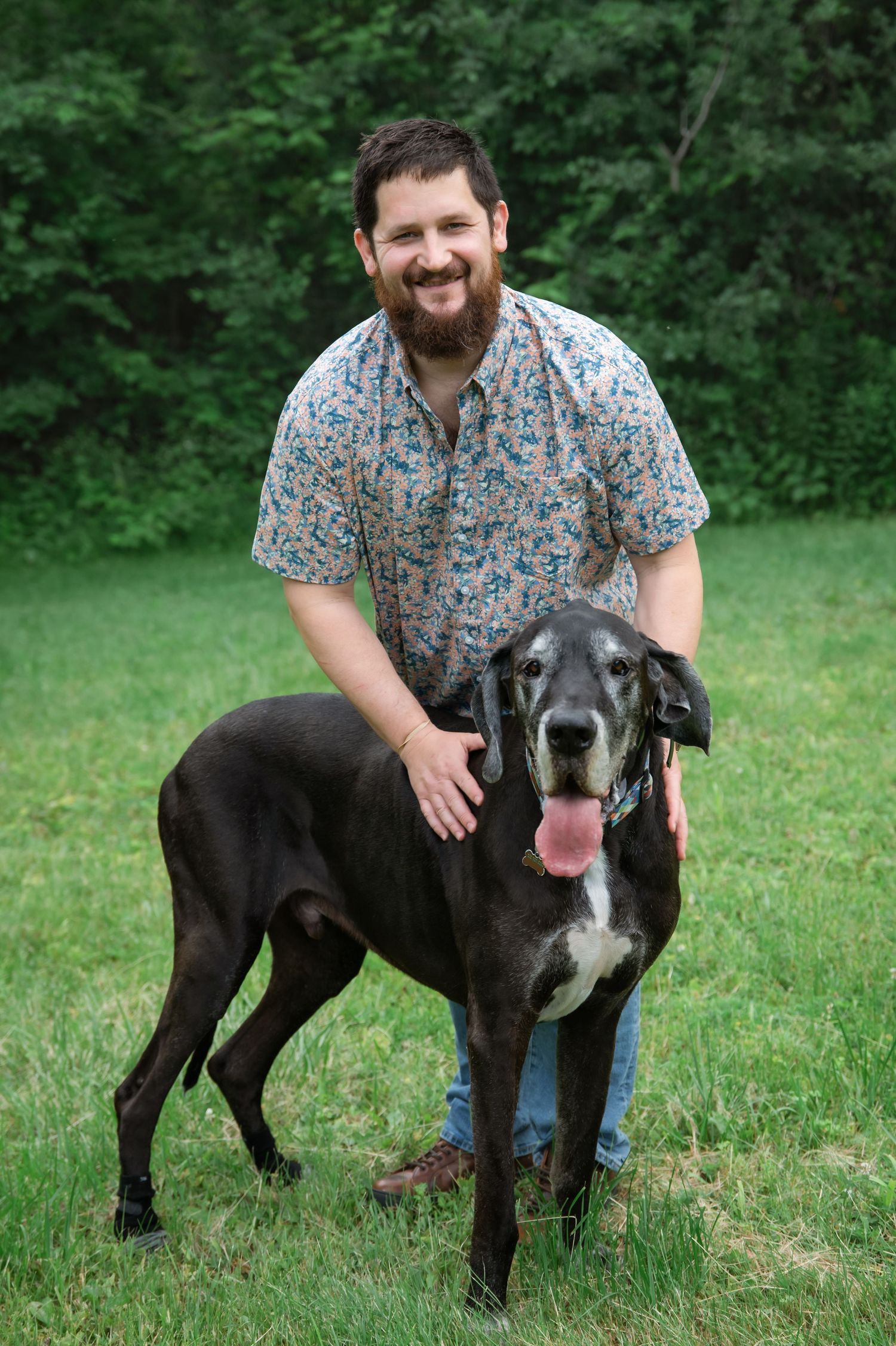 Smiling bearded man in floral shirt posing outdoors with a large black Great Dane dog on green grass.
