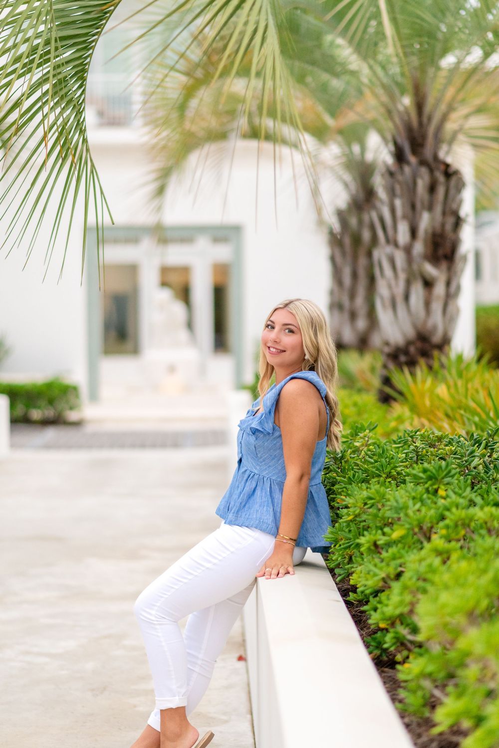 Smiling blonde woman in blue ruffled top and white jeans leaning on wall near tropical palm trees.