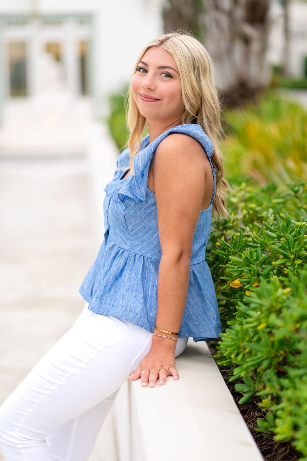 Young blonde woman in blue ruffle top and white jeans leaning on a wall with green shrubs outdoors.