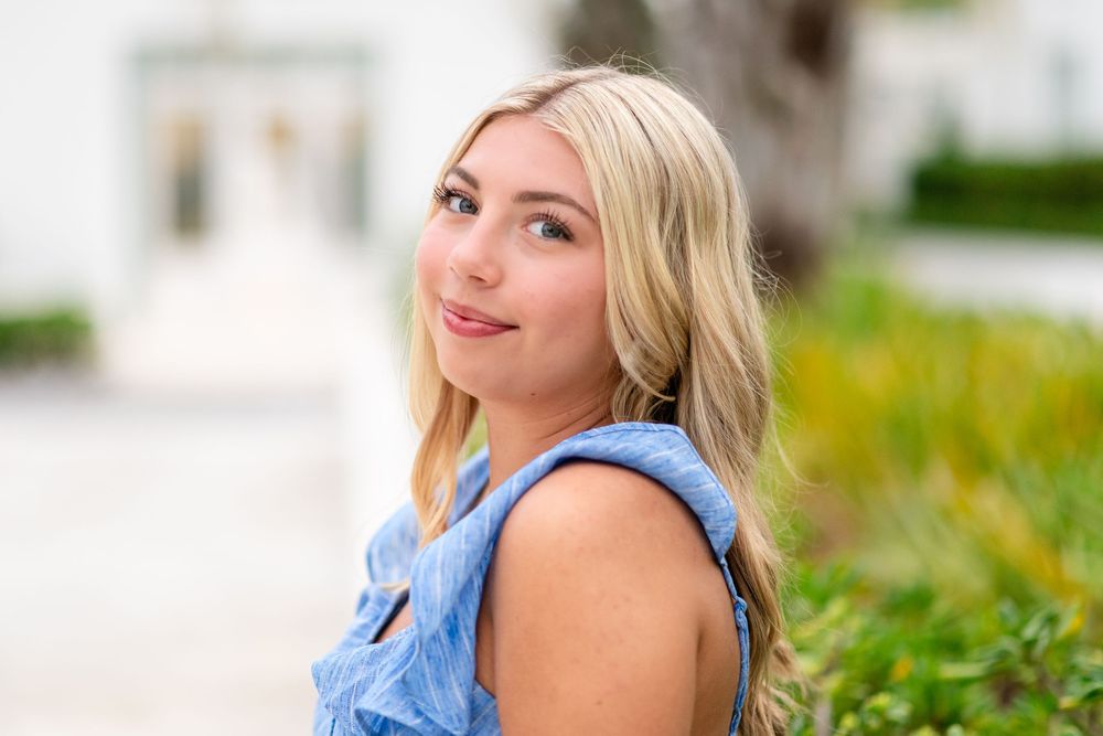 Young blonde woman in blue sleeveless top smiling over shoulder outdoors with green foliage background.