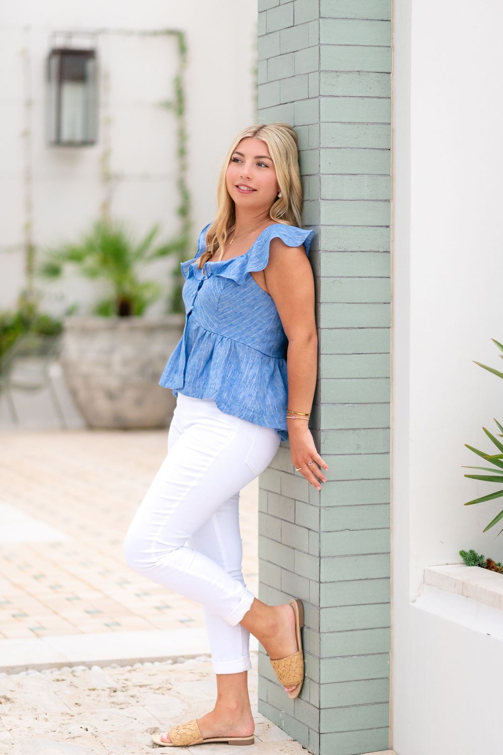 Blonde woman in blue ruffled sleeveless top and white jeans leaning against green tiled wall outdoors.