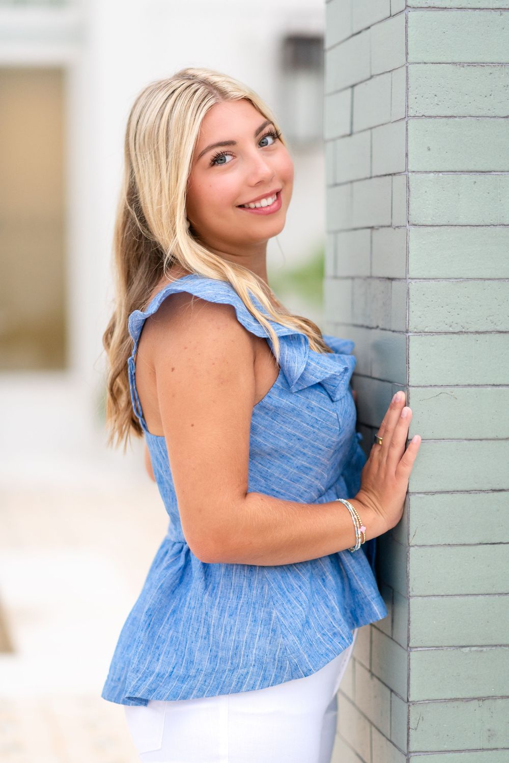 Smiling blonde woman in blue ruffled top leaning against a teal brick wall outdoors.