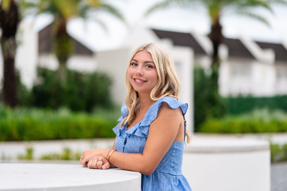 Smiling blonde woman in a blue ruffled dress leaning on a white railing outdoors with greenery and buildings behind her.