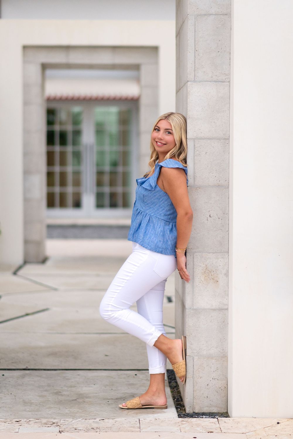 Smiling blonde woman in blue top and white jeans leaning against a stone pillar outdoors.