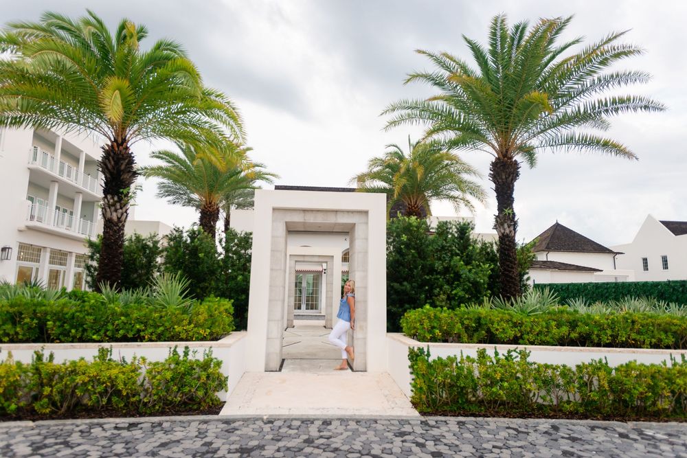 Woman walking through elegant white archway entrance flanked by tall palm trees at a luxury tropical resort.