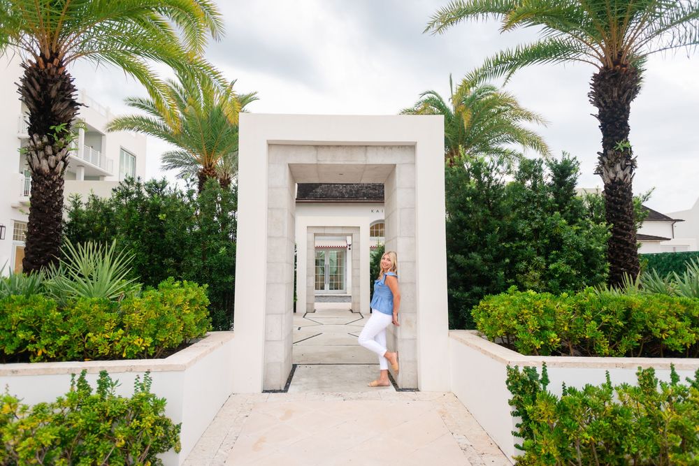 Woman in white pants poses by elegant stone archway entrance flanked by tropical palm trees at a luxury resort.