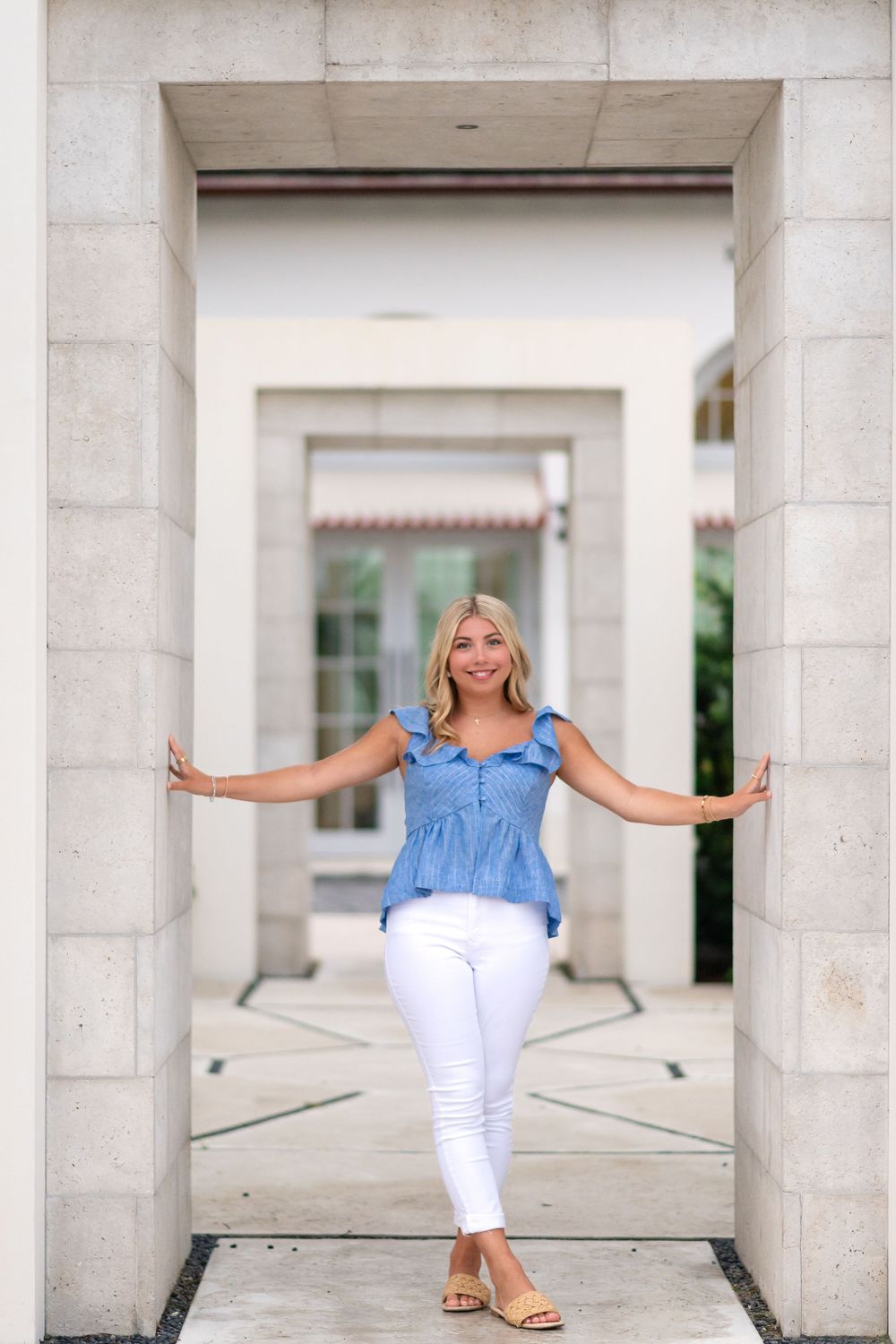 Blonde woman in blue top and white jeans posing in stone archway doorway.