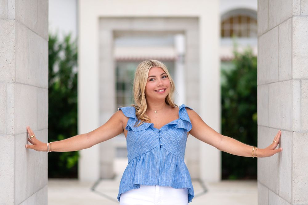 Smiling blonde woman in blue ruffled top posing between stone columns at an elegant outdoor venue.