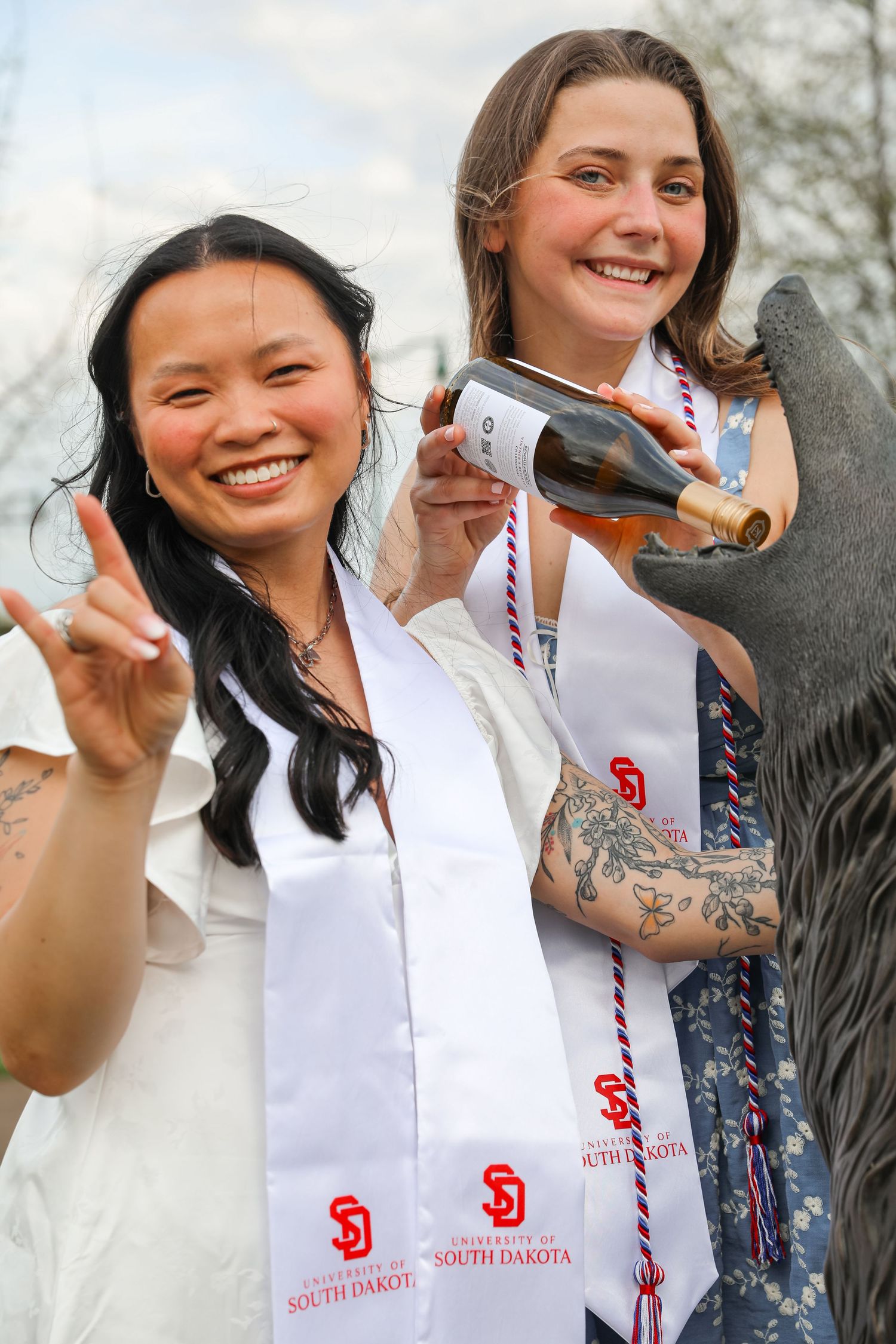 Two University of South Dakota graduates in white stoles smile and celebrate outdoors at commencement.