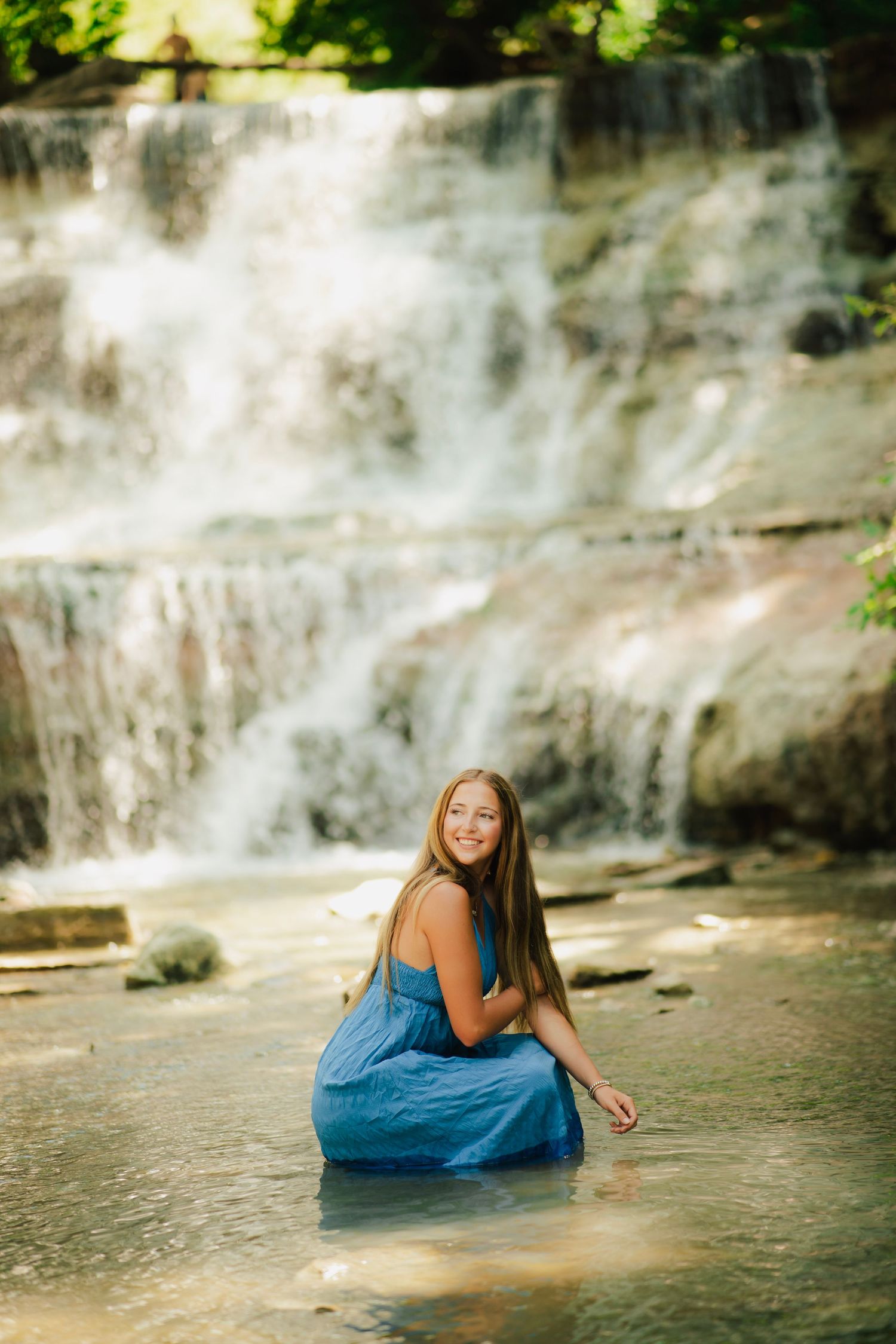 Young woman in blue dress sitting in shallow water before a cascading waterfall surrounded by lush greenery.