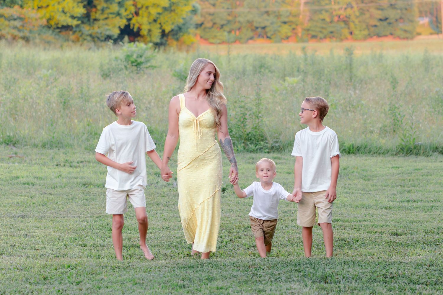 family photographer Calhoun, GA A mother in a yellow dress walks with her three children across a lush green field on a sunny day.