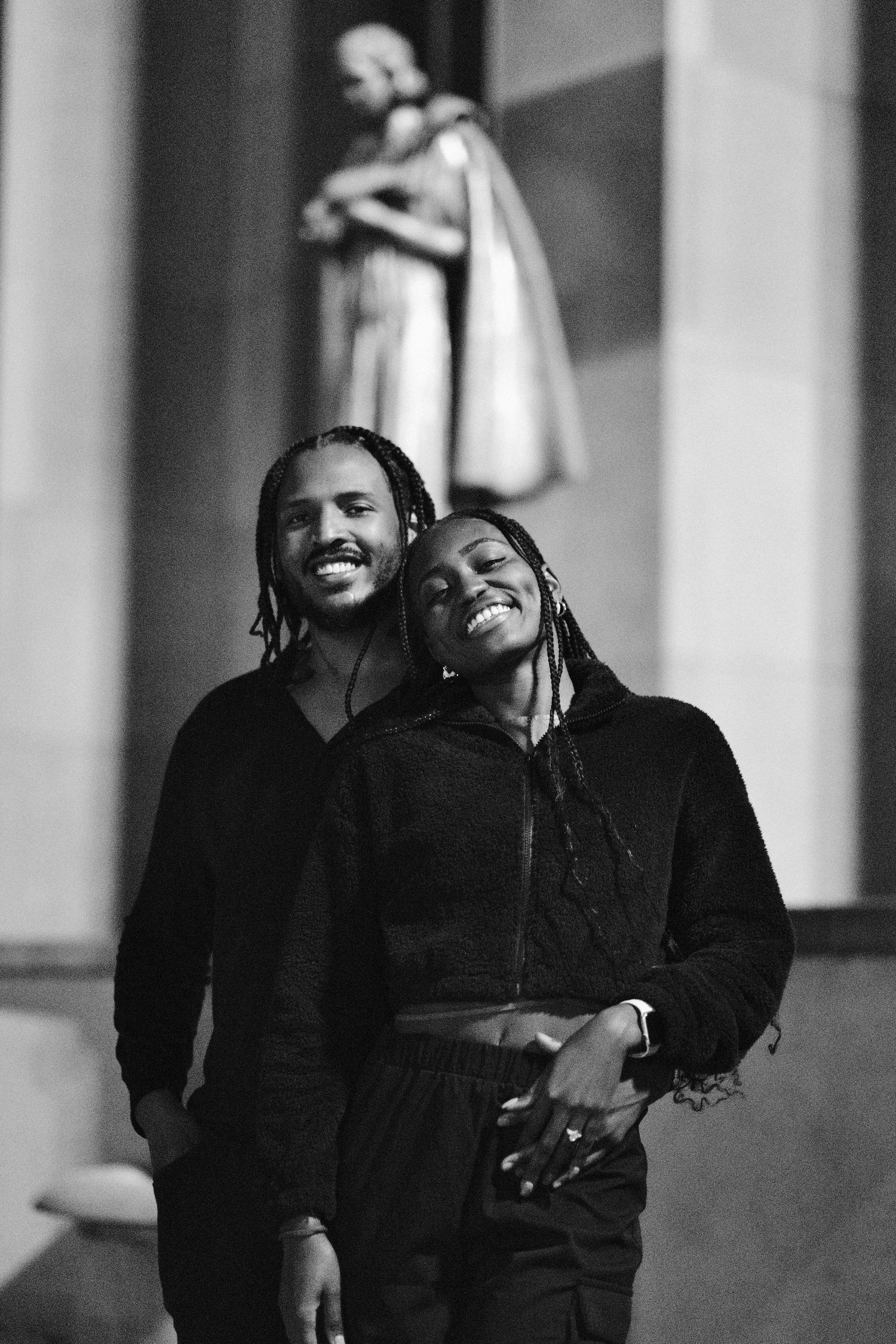 Black and white photo of two smiling people with braids embracing warmly in front of a classical building.