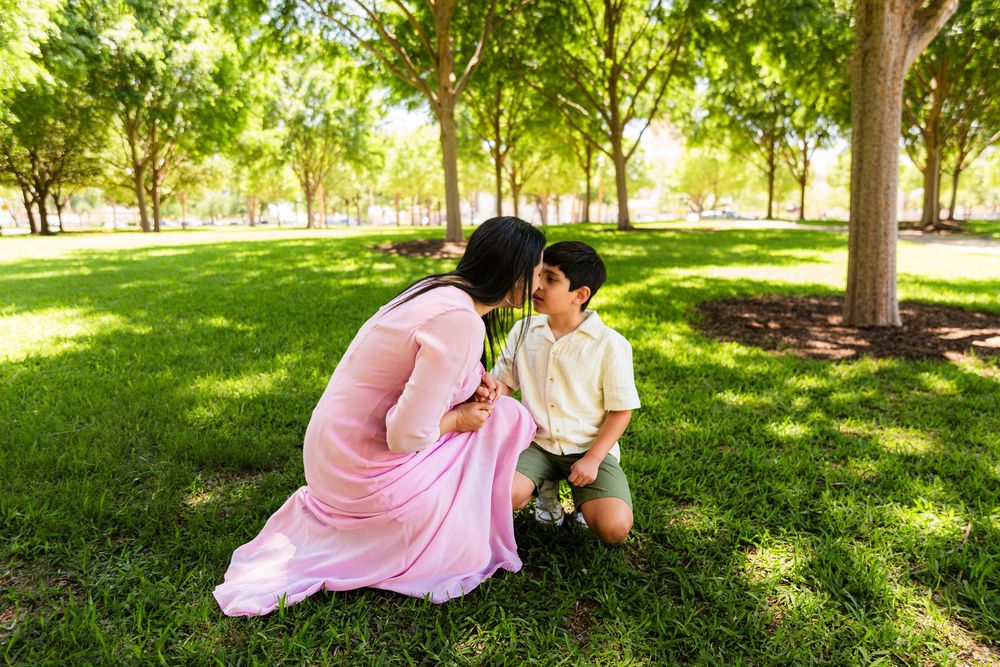 A mother in a pink sari kneels to kiss her young son in a lush green park surrounded by trees.