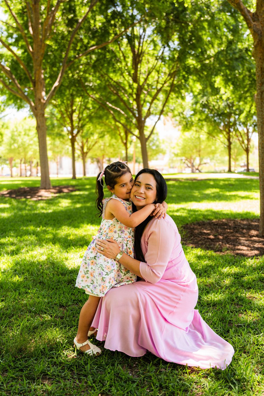 A mother in a pink dress hugs her young daughter in a floral dress outdoors in a lush green park.