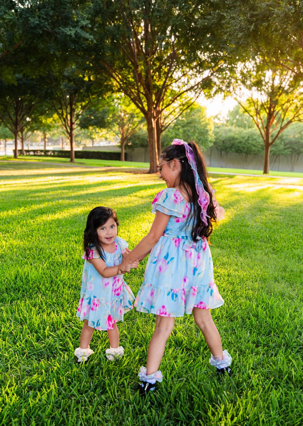 Two young girls in matching floral dresses hold hands and dance together on lush green grass in a sunny park.