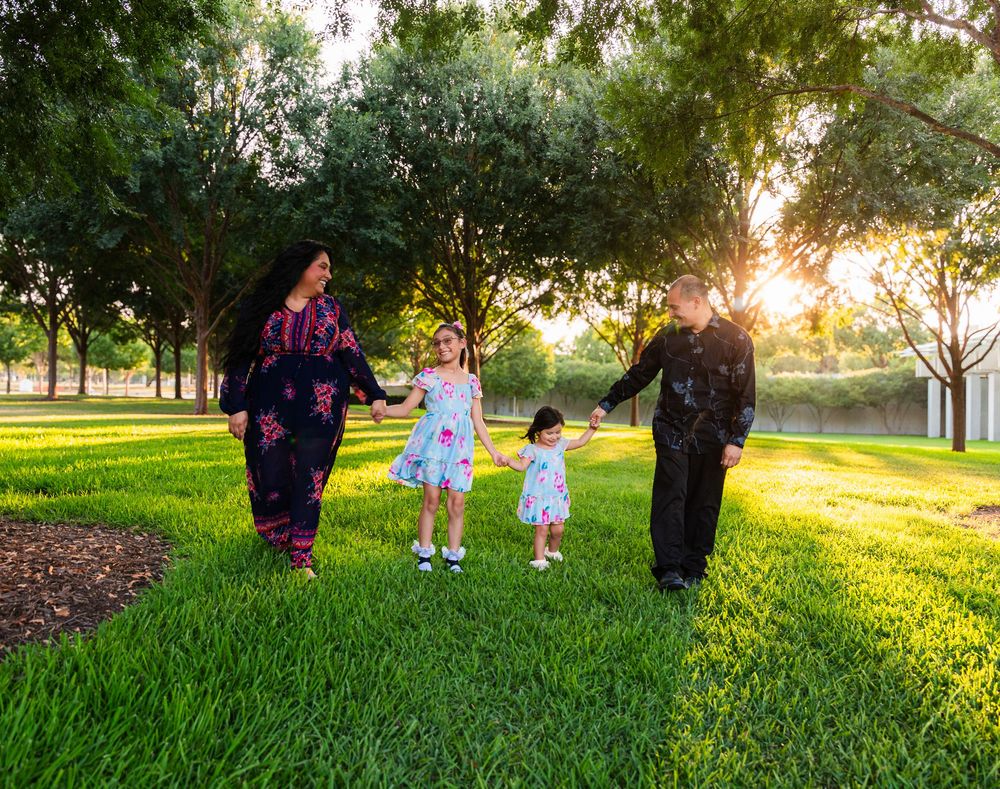 Family of four walking hand-in-hand through a sunlit green park at golden hour.
