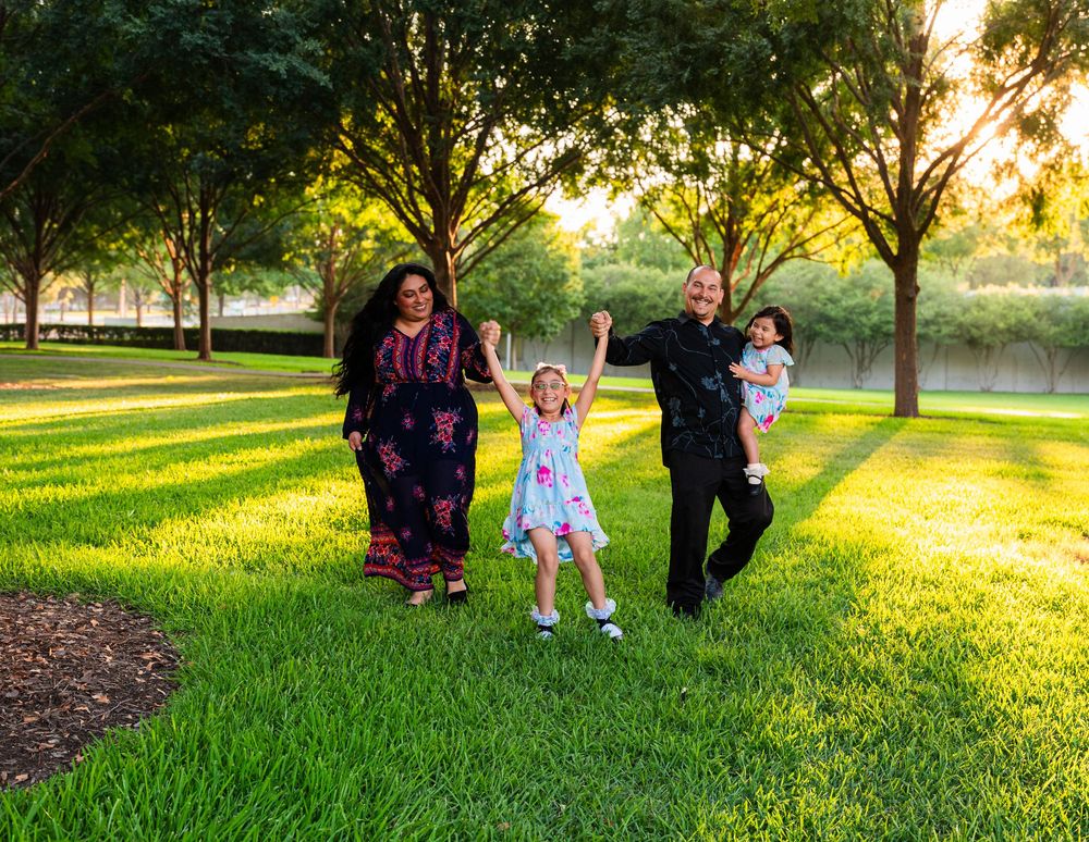 Family of four enjoying a sunny day in a lush green park, with two adults swinging a young girl between them.