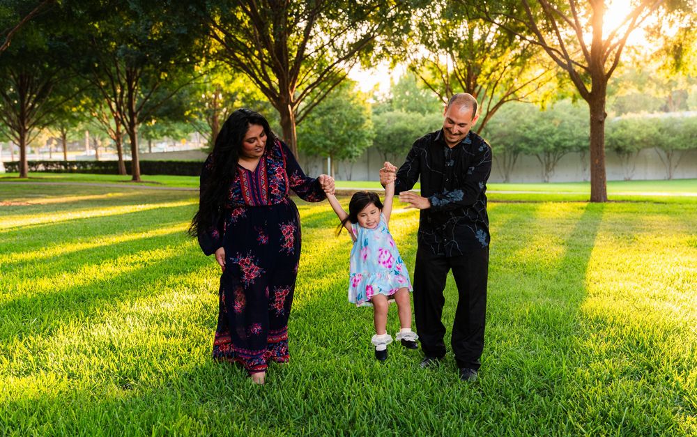 Family of three enjoying a sunny day in a lush green park, parents swinging their toddler daughter between them.