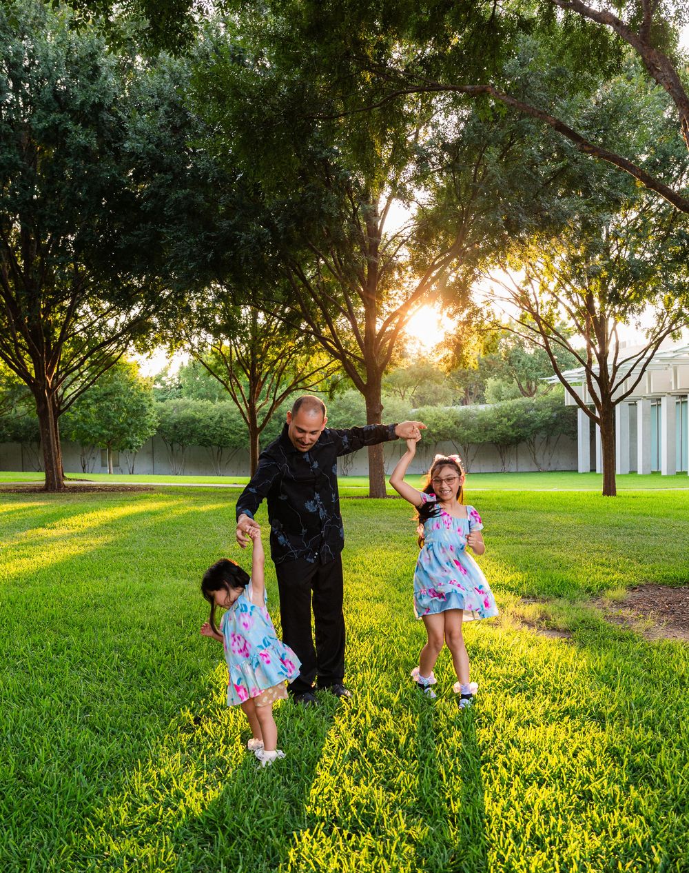 A father dances with two young daughters in a lush green park at golden sunset hour.