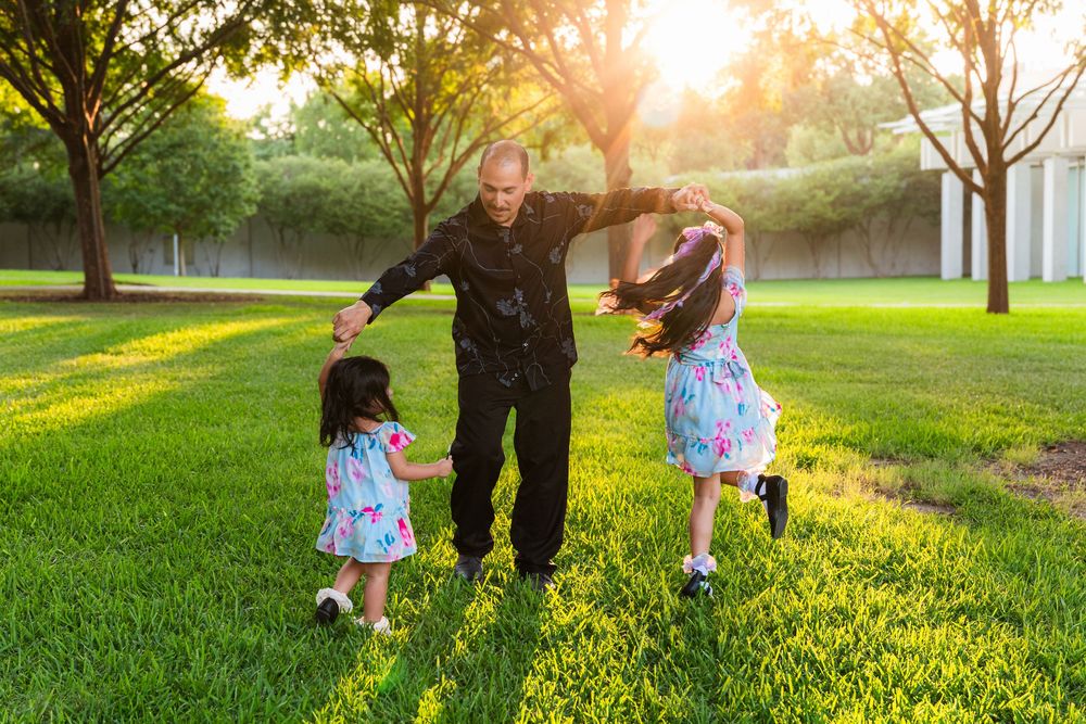 Man dancing outdoors with two young girls in floral dresses on green grass at sunset.